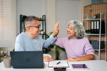 Asian Senior Couple High Five Celebrating Success with Laptop and Documents at Home Office Celebrating Retirement Planning and Financial Goals Achievement