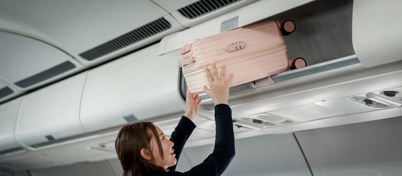 Happy  traveler stowing personal pink suitcase securely inside airplane cabin overhead compartment during boarding for business trip vacation journey