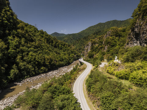 Aerial view of a winding road cutting through lush, green mountains alongside a river, creating a striking contrast with the bright sky, Khichauri, Adjara, Georgia.