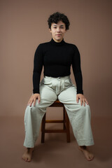 Young white woman with short curly hair, sitting on a stool, with hands on knees. Full body length studio portrait wth copy space on brown background. Ideal for Gen Z, individuality, self confidence.