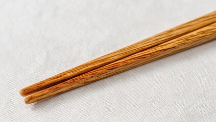 Close-up of a pair of light brown wooden chopsticks, slightly tapered, resting on an off-white textured fabric.  The wood grain is visible