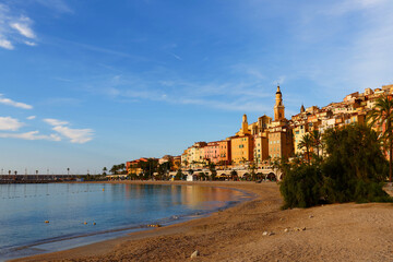 The sunset view of Menton, France, with colorful old town buildings glowing in warm light, set against the Mediterranean Sea and blue sky on the French Riviera
