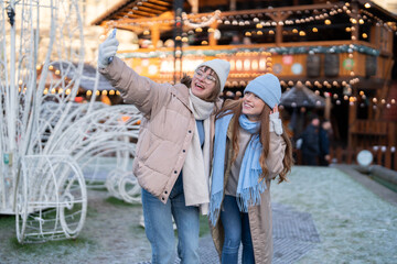 Friends enjoy winter day at local market taking selfie near festive decorations and lights in a cozy atmosphere during the holiday season