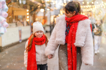 Cheerful woman and girl enjoy winter stroll at festive market with lights and snow in a cozy, holiday atmosphere during a snowy afternoon