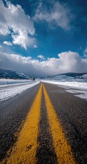 A snow-dusted asphalt road stretches to a vibrant blue sky, punctuated by fluffy white clouds.  Bright yellow double lines mark the center, vanishing into the distance