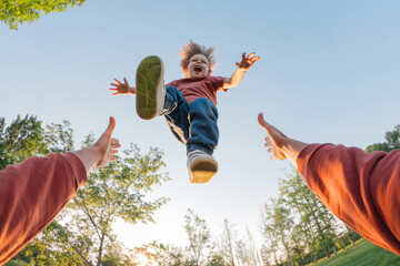 Amazed funny boy laughing while flies into the arms of his dad who tosses him high during a family fun time in a park. POV view