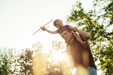 A father carries his excited son on his shoulders in a park, the boy holding a toy airplane and imagines flying as they enjoy a fun, carefree moment together. Copy space