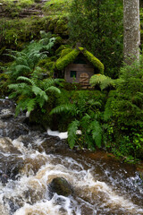 The image shows a small waterfall scene near the ruins of the Ieriķi Mill, where a stream flows through a lush green forest.