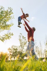 Joyful father tossing young son into the air, playing in a sunny park, surrounded by trees and fresh green grass at sunset. Fathers day and family bonding concept. Copy space