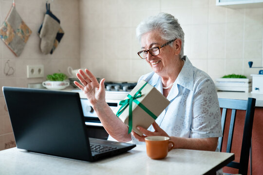 Happy senior woman holding gift box, talking online via laptop at home kitchen, having fun. Virtual celebration Thanksgiving, video call, holiday greeting, family connection. Technology lifestyle