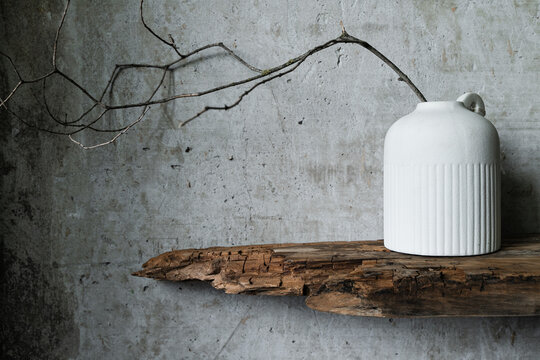 Minimalist still life with a white ceramic vase holding dry branches, placed against a rustic wooden board and textured concrete wall. Natural wabi-sabi style composition.