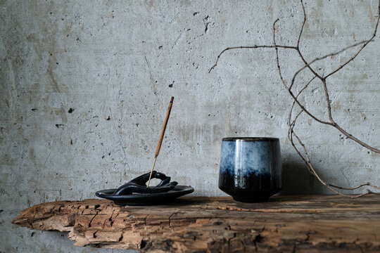 Burning incense stick in a hand-shaped holder with a ceramic cup on rustic wood against a concrete wall. Minimalist wabi-sabi still life.