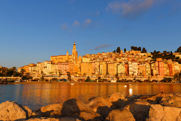 Panoramic sunset view of Menton, France, with colorful old town buildings glowing in warm light, set against the Mediterranean Sea and blue sky on the French Riviera