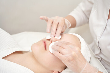 Caucasian female receiving facial treatment with cotton pads and gloves in spa setting.
