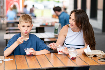 Cheerful mom and teen son laughing and eating ice cream at summer street cafe