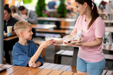 Mother handing ice cream to teenage son at outdoor summer cafe, warm family moment