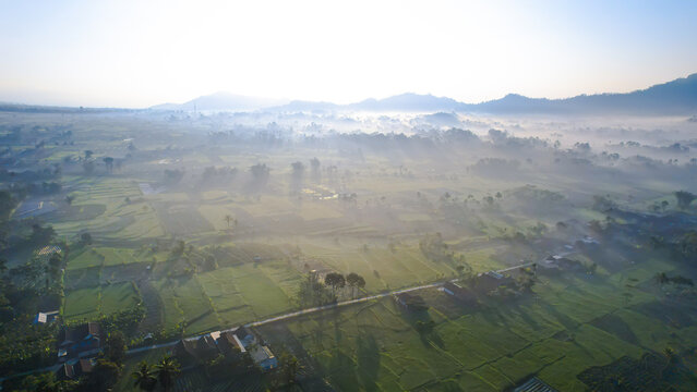 Aerial view from Teras Semeru shows green fields, scattered houses, and misty mountains under a clear sky, capturing the serene beauty around Mount Semeru.
