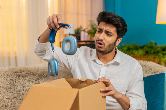 Sad Indian young man at home opening a package, shocked to find damaged wireless headphones inside. Arabian guy sitting on sofa, feeling disappointed and frustrated after receiving a broken order. - Powered by Adobe