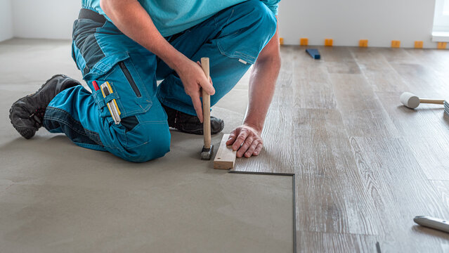 Floating floor work. The worker taps the board of vinyl plank to lock the click system. He is using hammer and wooden plank.
