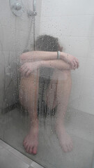 Depressed Person Sitting Alone on Shower Floor Behind Wet Glass with Head Down and Arms Wrapped...
