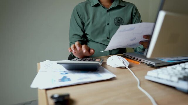 Person in a green shirt working at a desk with papers, calculator, and laptop