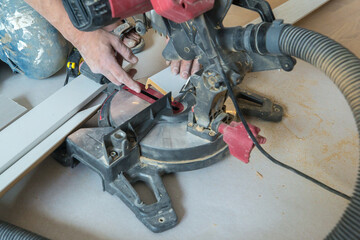 Close-up of worker using miter saw to cut white wooden board. Man's hands are visible holding the...