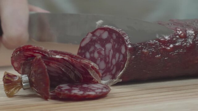Close-up of woman slicing smoked salami sausage into flat pieces on a cutting board, recorded with ambient sound.