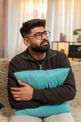 Indian man at home sits alone on sofa holding pillow, looking sad and emotionally distant. Arabian...