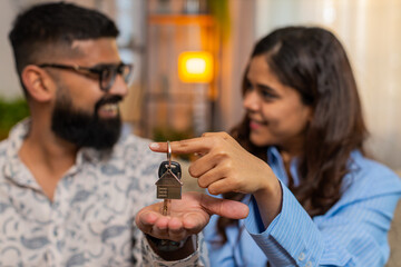 Indian couple man woman on home sofa, holding apartment keys and reading property contract with excitement and smiles. Young family celebrating successful new home purchase and happy agreement indoors