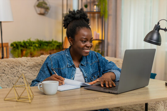African American young woman studies online at laptop while sitting at table on home sofa. Black girl takes notes in notebook looking focused, attentive, and fully engaged in distance learning process