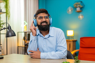Indian man at home sits happy expression wearing headphones sways gently dancing enjoying calm soft...