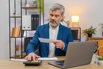 Middle-aged businessman at home office reviews budget today on laptop using calculator scanning...
