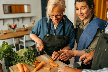 Smiling female chef assisting senior woman cutting carrots during cooking class in commercial kitchen