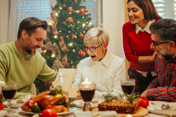 Family giving christmas gift to surprised senior woman during christmas dinner