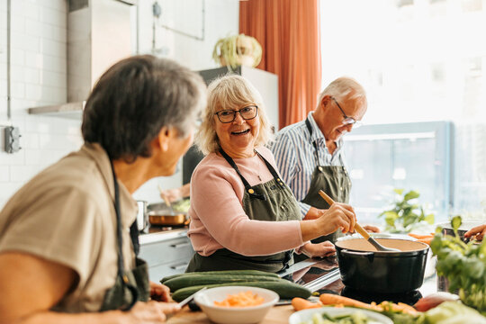 Happy senior woman stirring pot while talking with female friend during cooking class in commercial kitchen