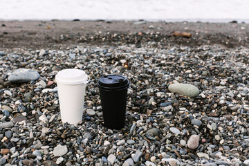 two disposable white and black coffee cardboard cup against the backdrop of the sea coast, sandy...