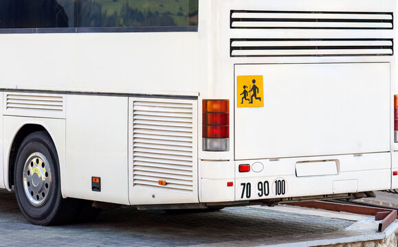 Rear view of a white coach bus with yellow school children warning sign, taillights and speed limit stickers 70 90 100, public transit vehicle parked at curb on city street, back part, kids transport