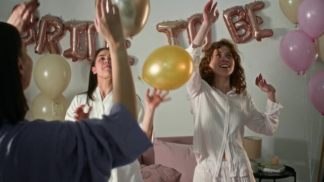 Happy girlfriends and bride-to-be in veil playing wit balloons, then hugging on bed while having fun during hen party