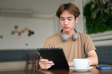 Young Thai student man in coffee shop wearing headphones and using digital tablet computer