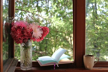 An open notebook, cup of coffee, and vase of bright autumn flowers on wooden windowsill. Sunlight...