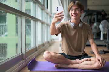 Young man sitting cross-legged on yoga mat using smartphone in gym