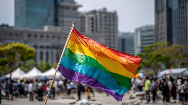 Colorful rainbow LGBT pride flag flying in urban setting with blurred crowd and modern city buildings during outdoor festival or parade event - Powered by Adobe