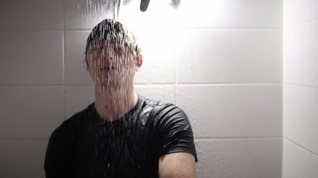 Teenager sits under shower streams with wet hair and dark shirt letting water cover his face creating a moody scene of reflection vulnerability and emotional atmosphere in the tiled space