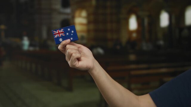 Hand of caucasian man holding small australian flag patch inside church with embroidery detail, highlighting cultural symbolism in indoor setting.