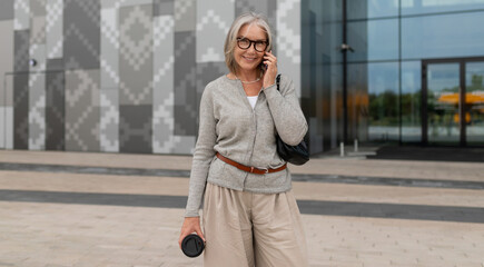 Senior woman talks on phone outside modern building while holding coffee in urban environment