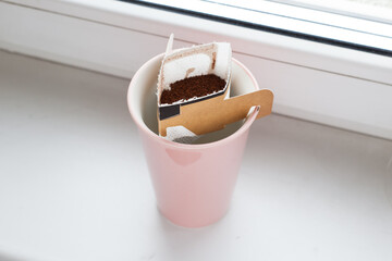 Drip bag in a ceramic cup on windowsill. Instant freshly brewed alternative filter drink in mug. Easy brew black coffee method for home, travel or office. cozy trendy lifestyle 