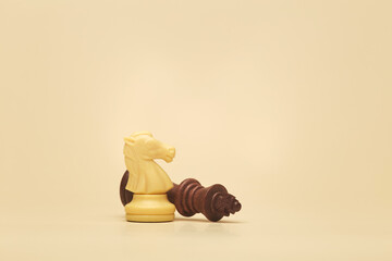 White chess knight standing next to a toppled brown king on beige background, symbolizing checkmate and victory in strategy © Antonio