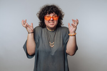 Person with curly hair wearing orange sunglasses and a gray shirt, gesturing with hands raised,...