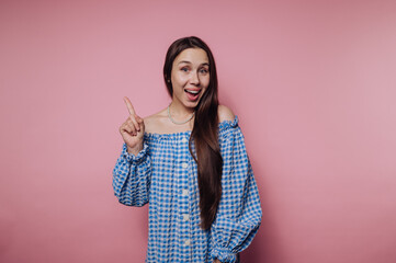 Smiling woman in blue checkered dress pointing up on pink background.