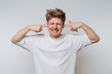 Young man in white shirt covering ears with fingers, eyes closed.